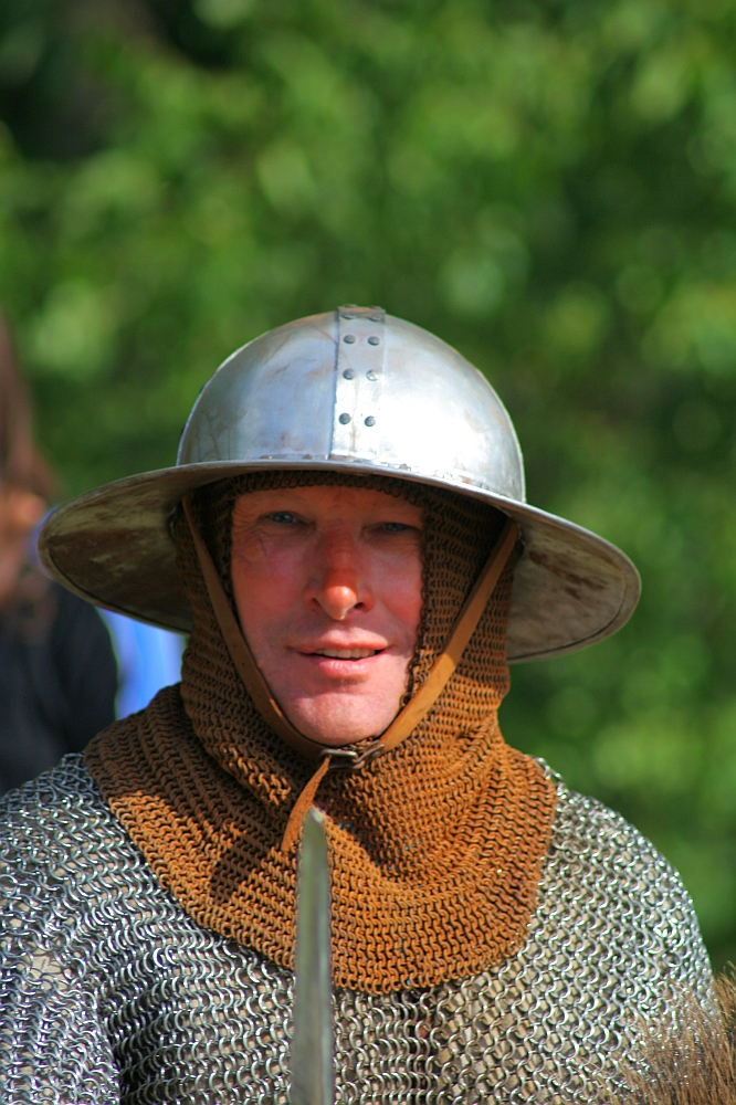 A person dressed in medieval armor, including a metal helmet and chainmail, looks directly at the camera. The individual has a faint smile and the background is blurred greenery, suggesting an outdoor setting during Semigallian days 2008. The chainmail hood is brown, indicating age or a specific design choice.