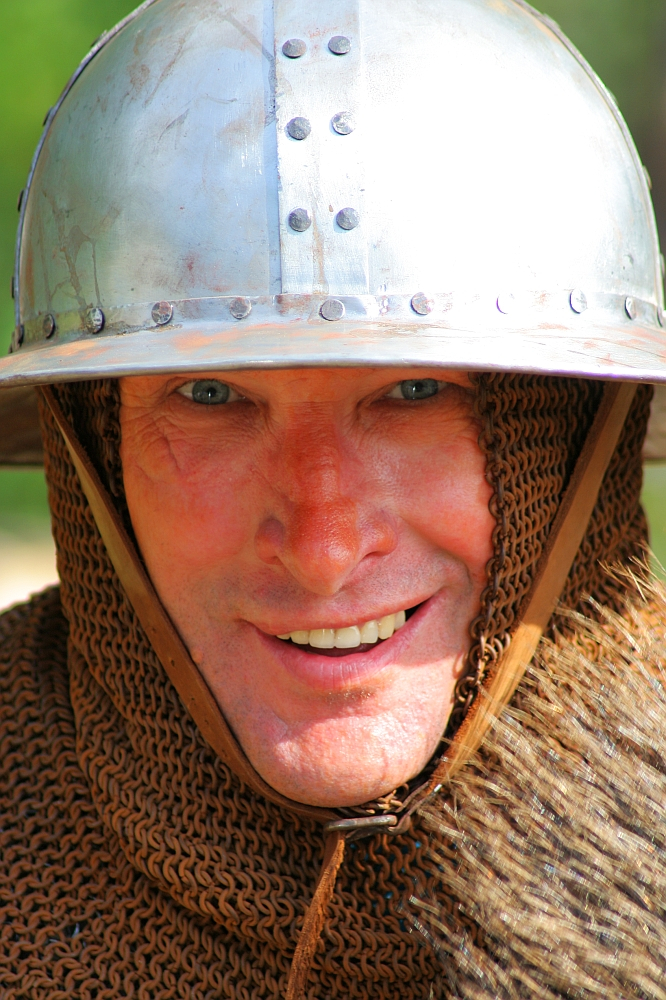 A person is smiling and wearing Semigallian medieval reenactment armor, which includes a metal helmet with rivets and a chainmail coif covering the head and neck. The background is blurry, emphasizing the individual’s detailed facial expression and historical attire. The lighting is bright and natural.