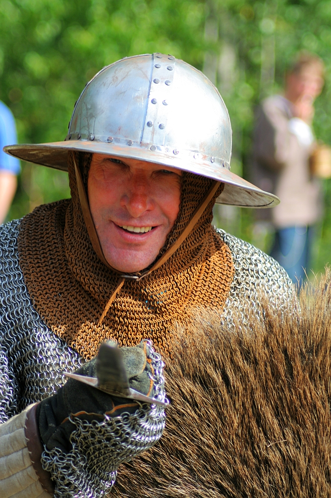 A person dressed in medieval chainmail armor and a metal helmet, smiling at the camera during Semigallian Days 2008. They have a chainmail coif under the helmet and are holding a fur-covered shield. The background is blurred, showing greenery and someone in modern clothing.