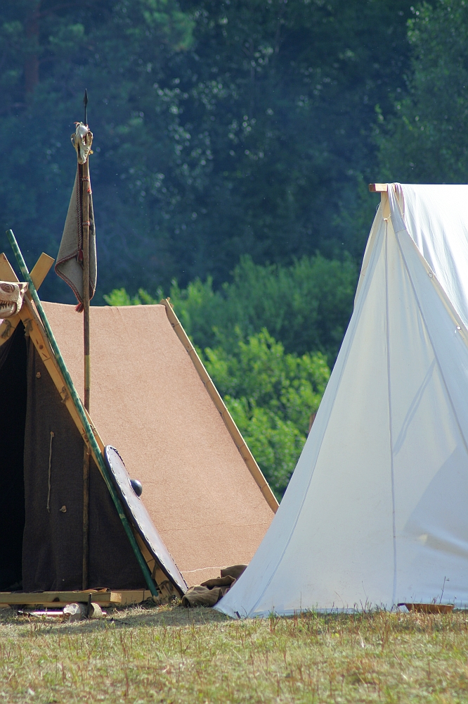 Two canvas tents are pitched in an open grassy area with a backdrop of dense, green trees, reminiscent of Semigallian Days. One tent is beige and constructed in a traditional triangular shape with a small opening. The other tent is white and has a larger and more structured appearance, evoking the spirit of the 2008 event. Various camping items are scattered around.