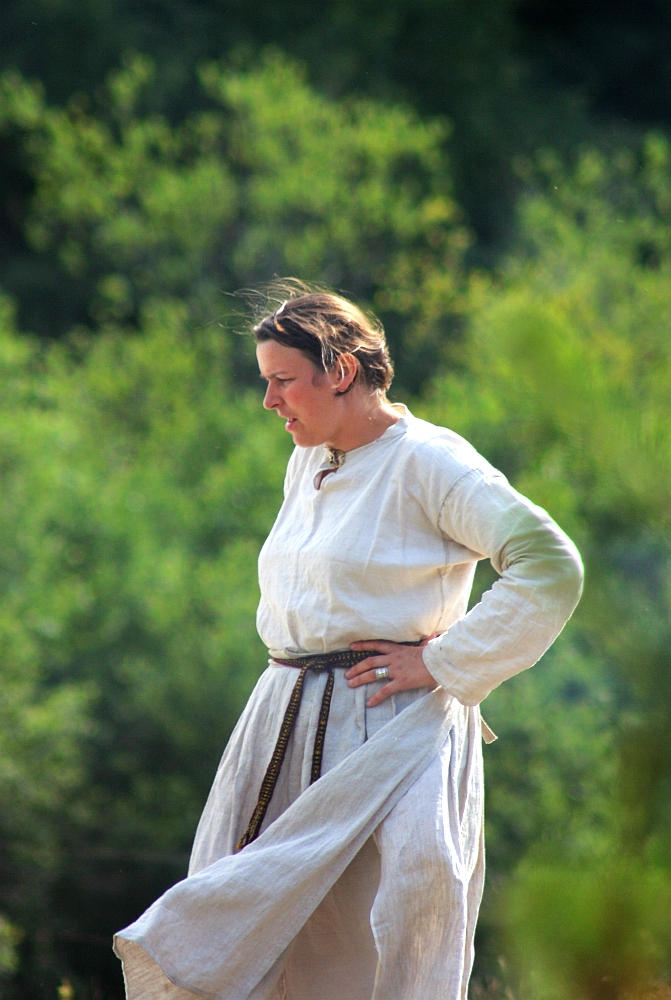A person is shown standing outdoors during the '08 Semigallian days festival, wearing a long, white garment with a brown belt. The individual has short, light-colored hair and appears to be looking down or to the side. The background is a blurred view of green foliage, suggesting a natural, outdoor setting.