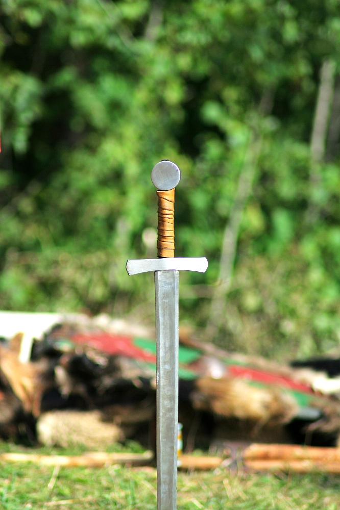 A medieval sword stands vertically embedded in the ground against a blurred green forest background. The sword, reminiscent of those from Semigallian days, features a straight cross guard, a round pommel, and a grip wrapped in brown leather.