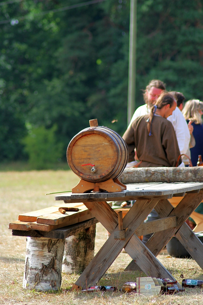 A small wooden barrel is placed on a rustic wooden table, set outdoors with a background of green trees. Several people are gathered near the table, engaged in conversation. A wooden bench and scattered bottles are visible in the foreground, evoking memories of relaxed Semigallian days and casual outdoor gatherings.