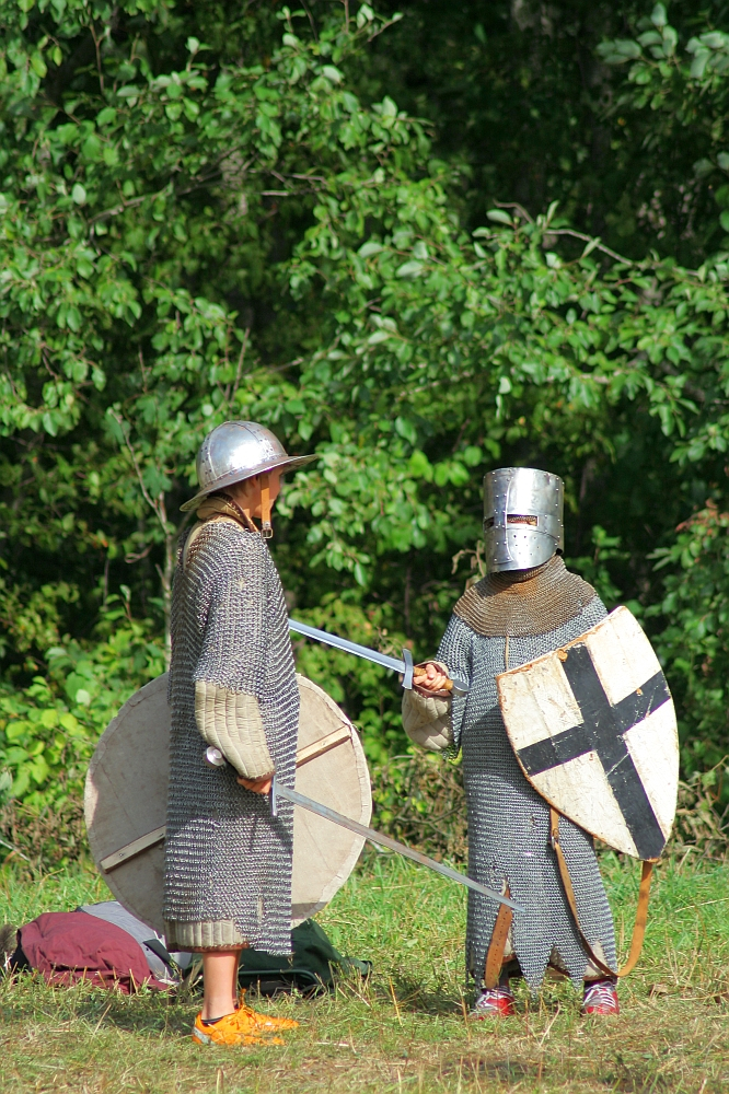 Two people dressed in medieval armor engage in a mock sword fight in a wooded area during the annual Semigallian Days event. They wear chainmail tunics and helmets. One holds a round shield, and the other has a shield with a black cross. Both hold swords, with a backpack on the ground behind them.