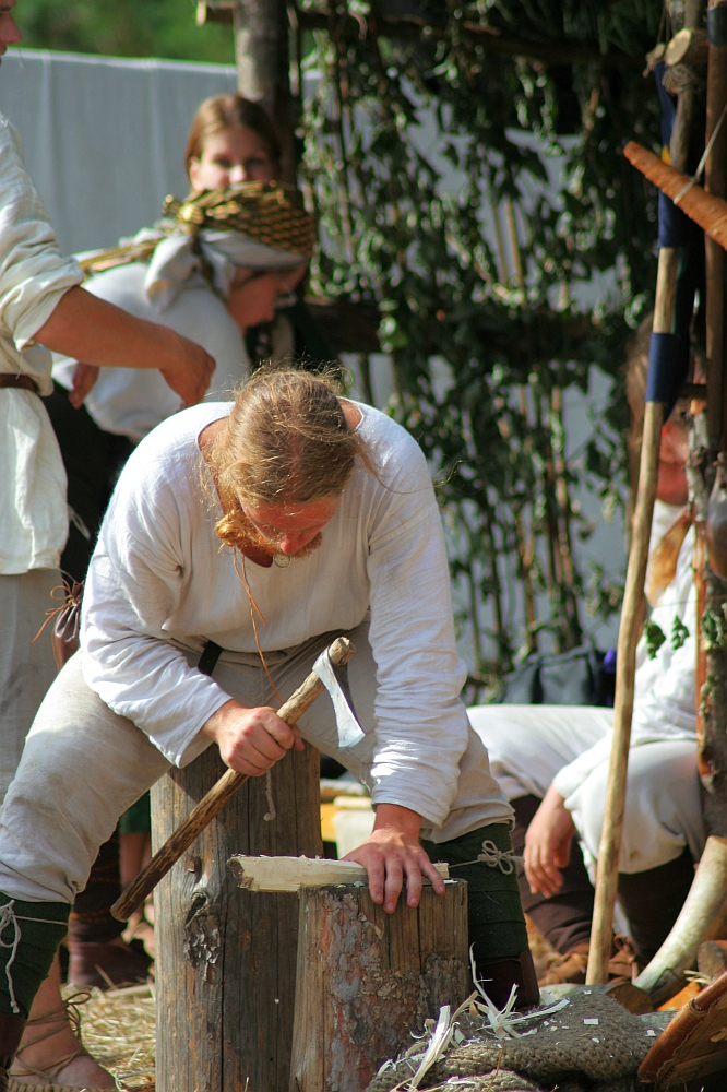 A person dressed in historical Semigallian clothing is chiseling wood while kneeling on the ground. Other individuals, also in period attire, are visible in the background. The setting appears to be a days-long outdoor reenactment camp with greenery and rustic wooden structures, reminiscent of 2008 events.