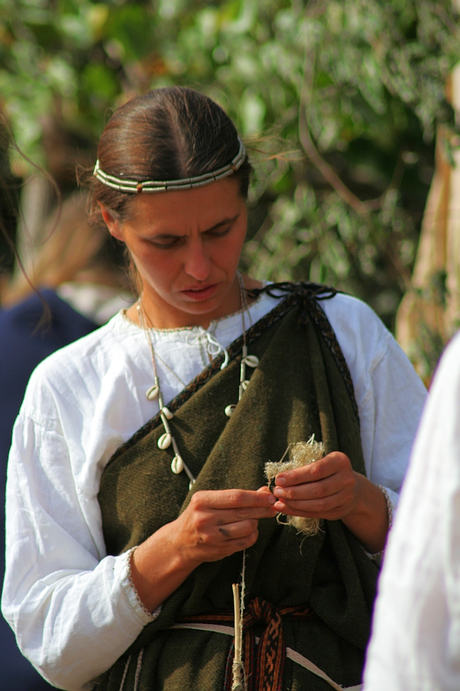 A woman with long brown hair, adorned in a simple headband, wearing a white blouse and a dark green draped garment, stands outdoors. She is focused on weaving or knotting a bundle of fibers in her hands during Semigallian Days 2008. She also wears a necklace made of light-colored beads. The background is blurred greenery.