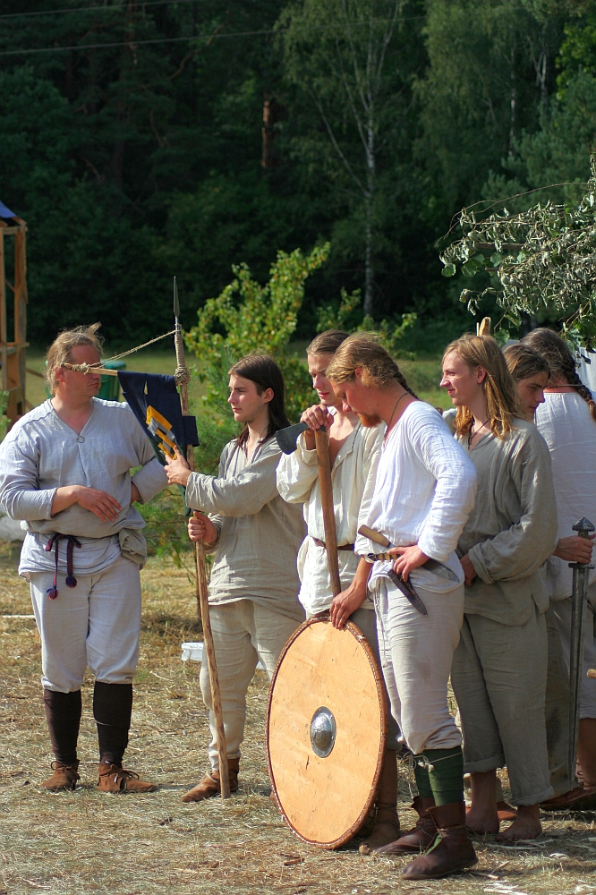 In a 2008 reenactment, a group of people dressed in historical medieval attire stands outdoors in a forest clearing. They are wearing white tunics and holding various props like wooden poles, a round shield, and a flag. One person appears to be speaking to the group. Trees and greenery are visible in the background, reminiscent of old Semigallian days.
