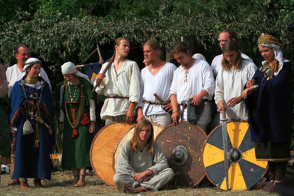 A group of people, dressed in historical Viking attire, pose outdoors as if transported from Semigallian days. Men and women stand behind three large round shields with colorful designs, and a young man sits on the ground in front. The background is lush green foliage and a structure made of branches. Some wear jewelry and ornate clothing.