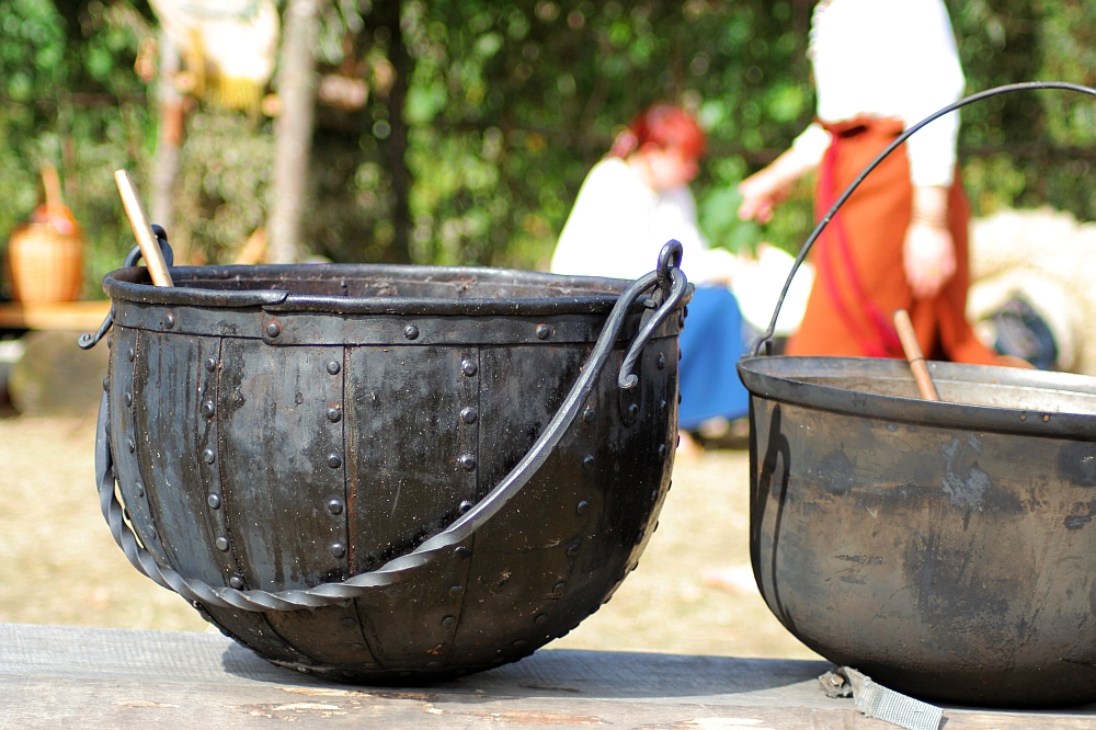 A large, black, cast-iron kettle with a handle and a stir stick rests on a wooden surface outdoors. In the blurred background, two people dressed in historical attire, including a white shirt and a red skirt, are engaged in conversation. This scene is from the Semigallian festival held in 2008, surrounded by lush greenery.