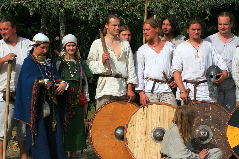 A group of people dressed in medieval attire are gathered together at a Semigallian festival. On the left, a woman wears a white headpiece and elaborate jewelry, while the men around her wear simple tunics. Some men hold round wooden shields and swords. The background features greenery and a rustic setup reminiscent of 2008 Semigallian days.