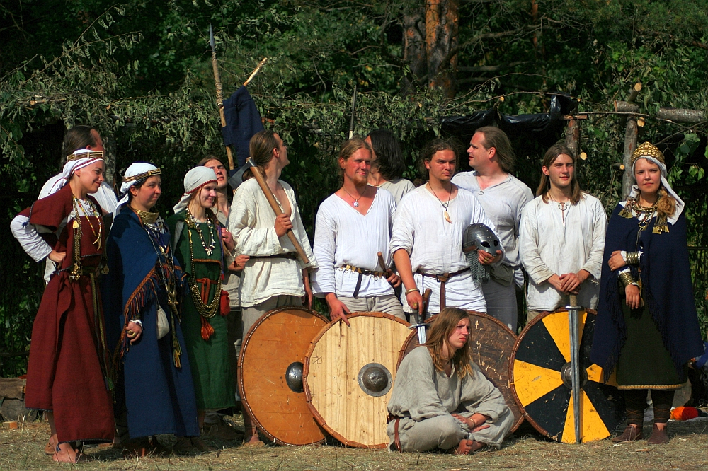 A group of eleven people in medieval clothing stands outdoors during the Semigallian Days. Some wear white tunics while others are dressed in blue and green robes adorned with jewelry. Three hold round wooden shields, and one person kneels in front with blonde hair. The background has green foliage and trees.