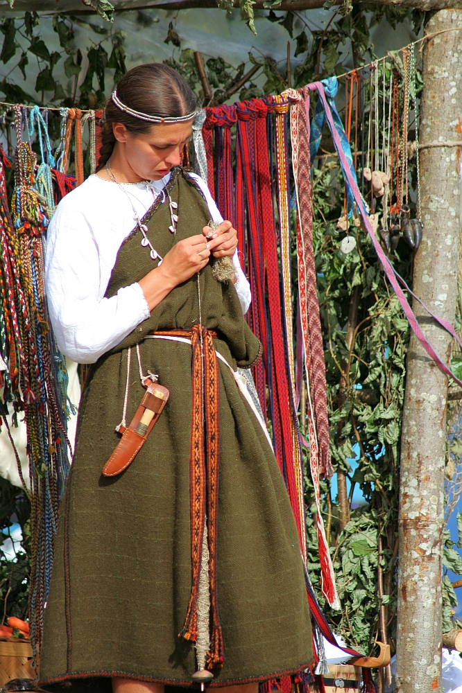 A woman wearing a medieval-style green dress and a white long-sleeve shirt is standing in front of an array of colorful ribbons hanging in the background. She is focused on her hands, possibly crafting or tying a knot. A brown belt with a sheathed knife hangs at her waist, reminiscent of 2008's Semigallian Days celebration.