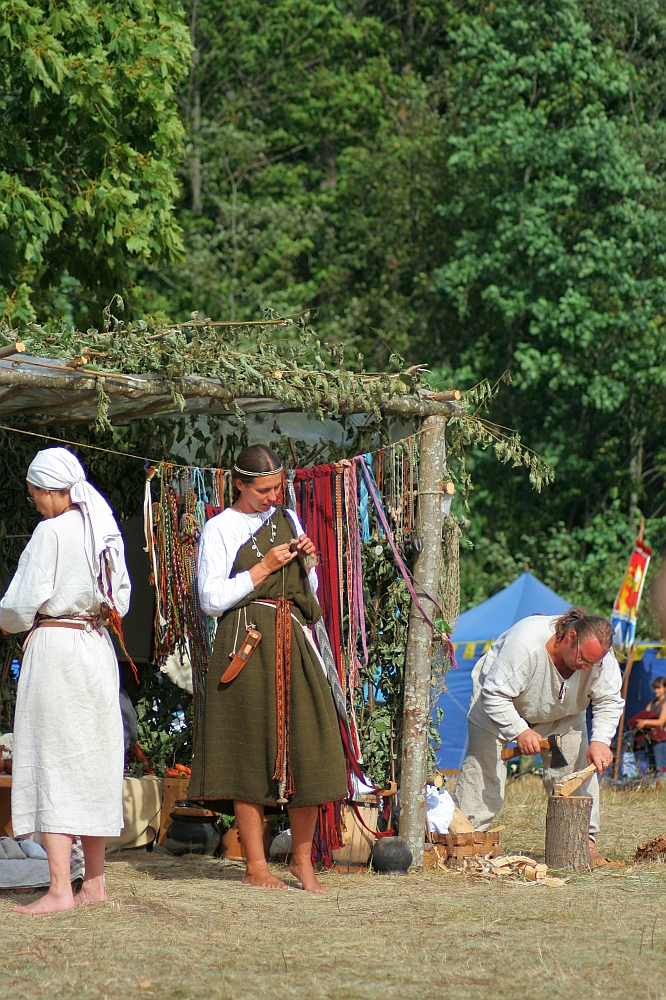 Three people dressed in historical clothing are partaking in an outdoor, medieval reenactment at Semigallian Days '08. One person stands under a canopy with colorful ribbons; another crafts an item on the right, while a third, seen from the back, is to the left. Trees and a tent are visible in the background.
