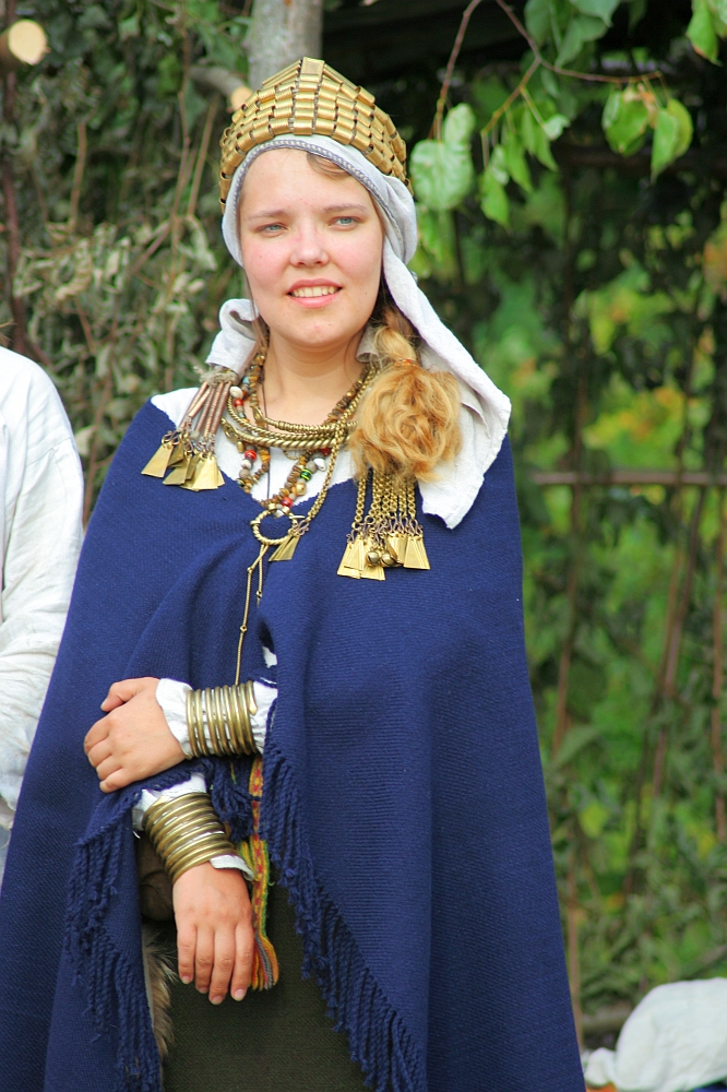 A person dressed in traditional attire stands outdoors during Semigallian Days 2008. They are wearing a blue cloak, several necklaces with large pendants, and arm bracelets. A white headscarf with a golden headdress adorns their head while green foliage is visible in the background.