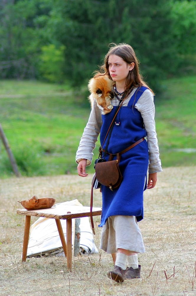 A young person wearing historical Semigallian attire, including a blue tunic over a beige shirt and beige pants, stands outdoors on a dry grassy area reminiscent of days past. They have a brown leather bag and a small fur accessory. A simple wooden table and bench with wooden bowls and a spoon are nearby, with green trees in the background.