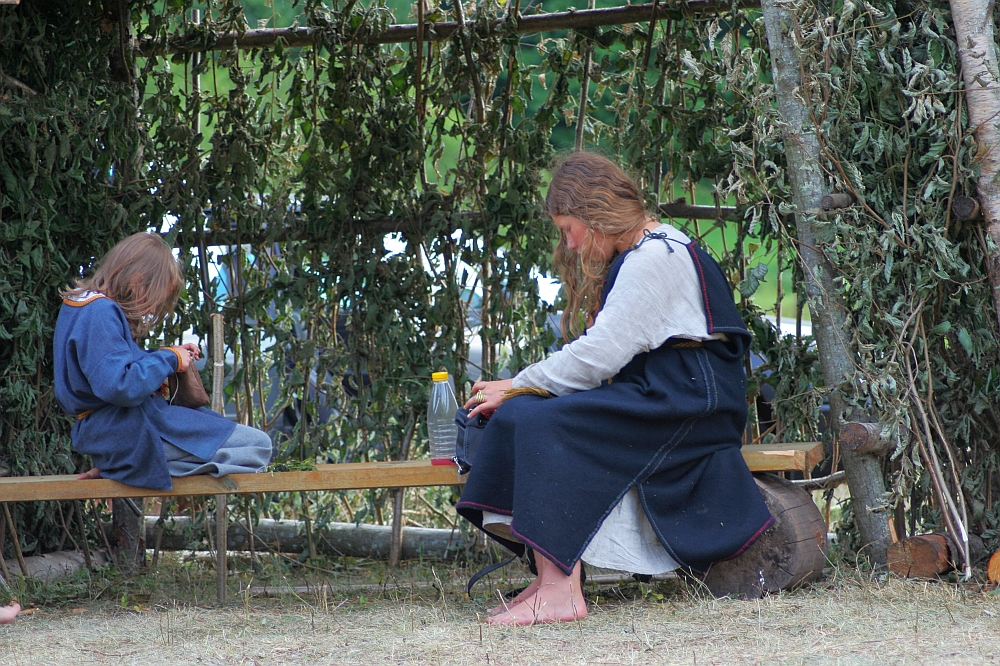 A woman and a child dressed in traditional medieval clothes sit on a wooden bench under a canopy of leafy branches. The woman is occupied with a task, while the child is engrossed in another activity. This rustic, natural setting harks back to the ambiance of Semigallian Days '08, surrounded by greenery.