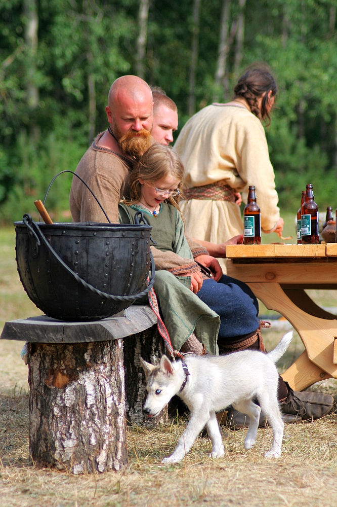 A bearded man sits on a log bench with a young child on his lap near a large black pot, reminiscent of the lively 2008 event, Semigallian Days. Next to them are two men seated at a wooden picnic table with bottles. A small white puppy on a leash stands in the foreground of the outdoor, wooded area.