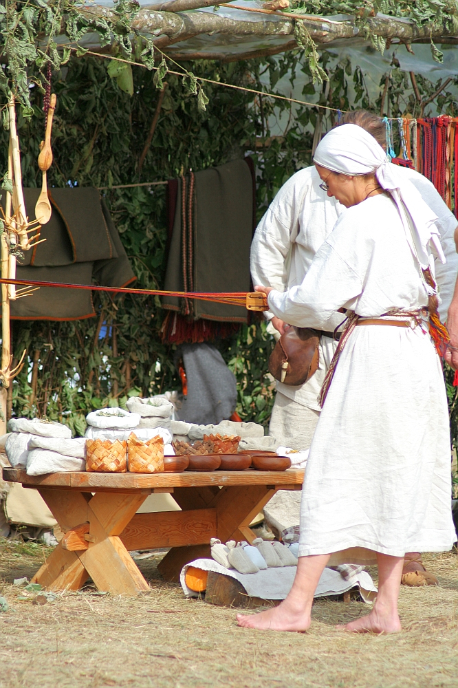 At Semigallian Days 2008, a person in a white tunic and headscarf, wielding a weaving tool with red threads, stands near a wooden table filled with baskets and bowls. Behind them, hanging fabric and wooden utensils adorn the structure, evoking an outdoor market or fair.