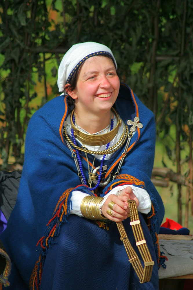 A smiling woman wearing a white headscarf and a royal blue cloak adorned with colorful tassels sits in front of a greenery backdrop. She wears various necklaces and bracelets, holding a small wooden instrument in her hands, reminiscent of the vibrant Semigallian days. She appears relaxed and content.