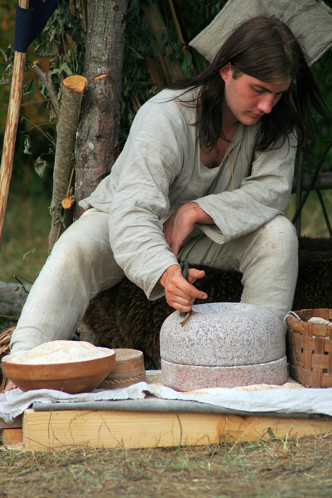 A man with long brown hair, dressed in a light-colored medieval-style tunic and pants, uses a small hand tool to grind grain on a stone mill. Surrounding him are wooden bowls filled with grain and flour, and a woven basket. The scene evokes the historical charm of Semigallian days circa 2008 amidst outdoor greenery.