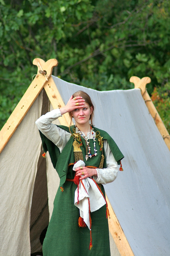 During the 2008 Semigallian Days, a woman stands in front of a canvas tent, wearing a green dress adorned with various accessories. She has her right hand raised to her forehead, partially covering her eyes. The background features green foliage, suggesting an outdoor setting. She holds a white cloth with red edges in her left hand.