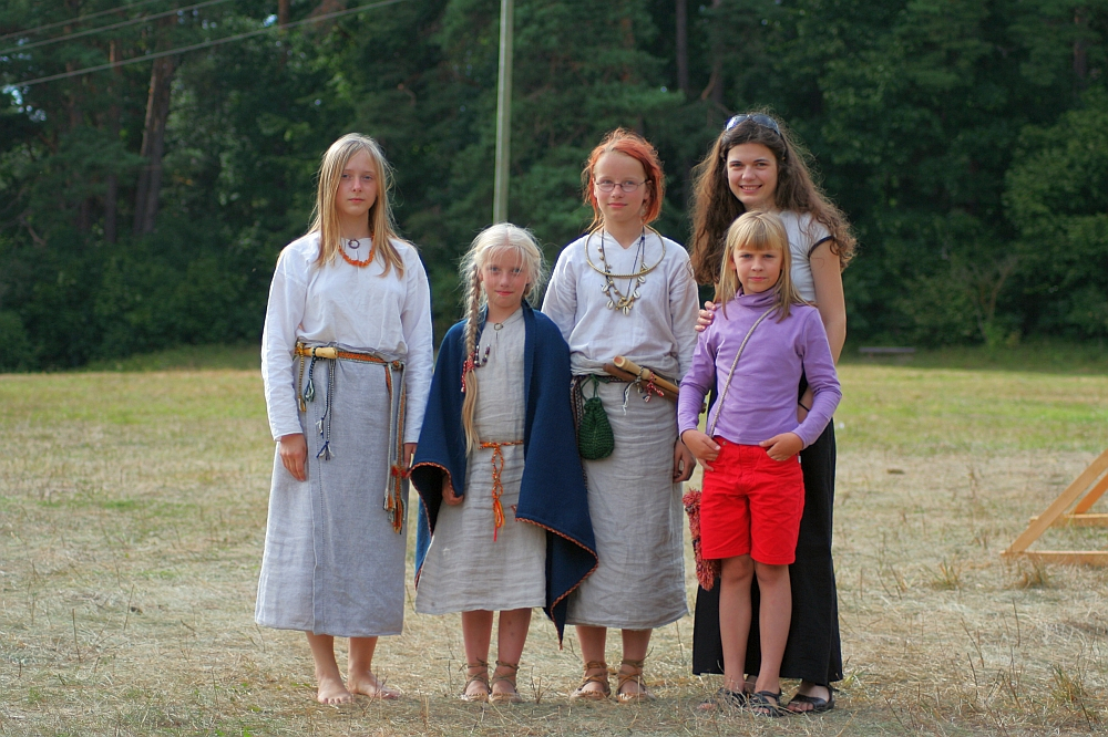 Five people stand outdoors in front of a forest background during Semigallian Days. Three women and two young girls, all wearing light-colored, loose-fitting garments from '08. One woman has a red bandana and beaded necklaces. One girl wears a purple top and red shorts. They are all barefoot on the grassy ground.