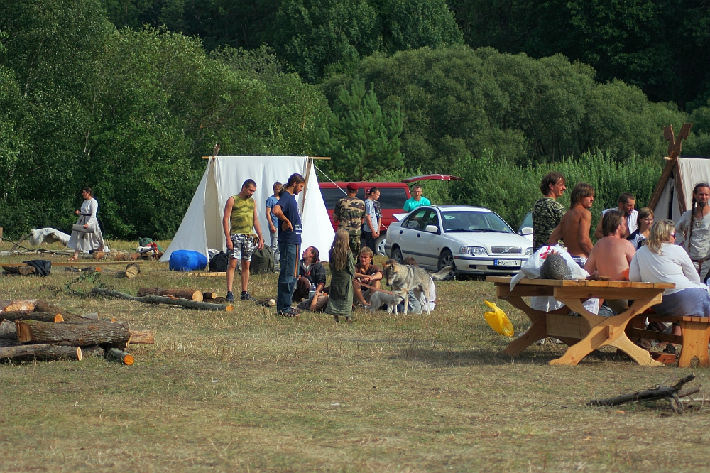 People are gathered at a campsite with white tents during the 2008 Semigallian Days event. Some are sitting at wooden picnic tables, while others are standing or sitting on the ground. A few dogs are present. Logs and camping gear are scattered around, and a silver car is parked nearby. The area is surrounded by trees and grass.