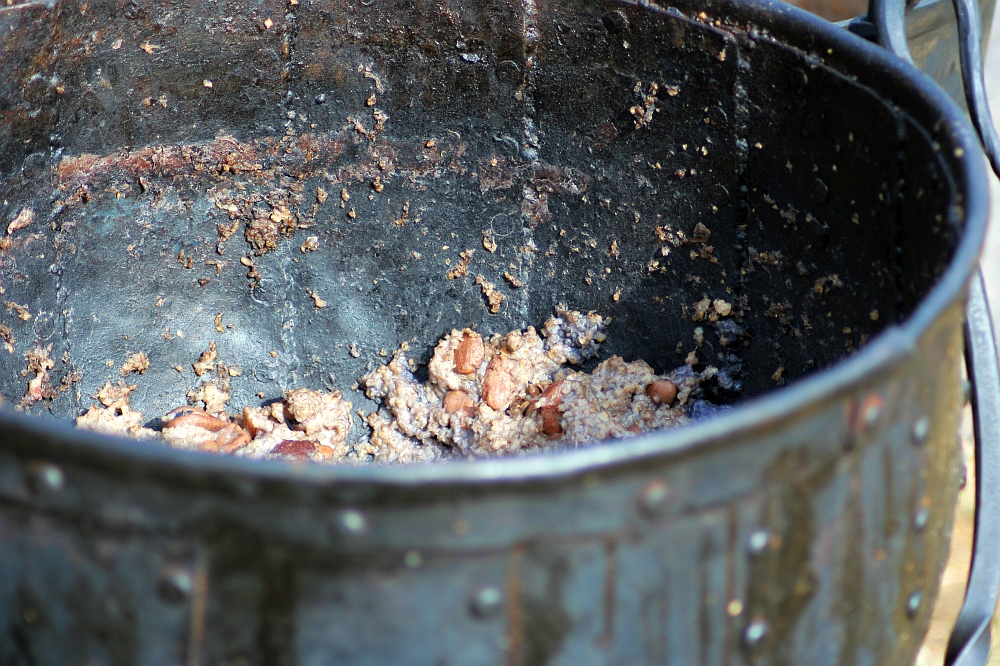 A large, aged metallic pot from the 2008 Semigallian Days event has a heavily worn interior containing remnants of cooked beans and grains. The food appears partially eaten, and the interior surface shows significant discoloration and texture from use. The pot is round, with visible rivets along the exterior.