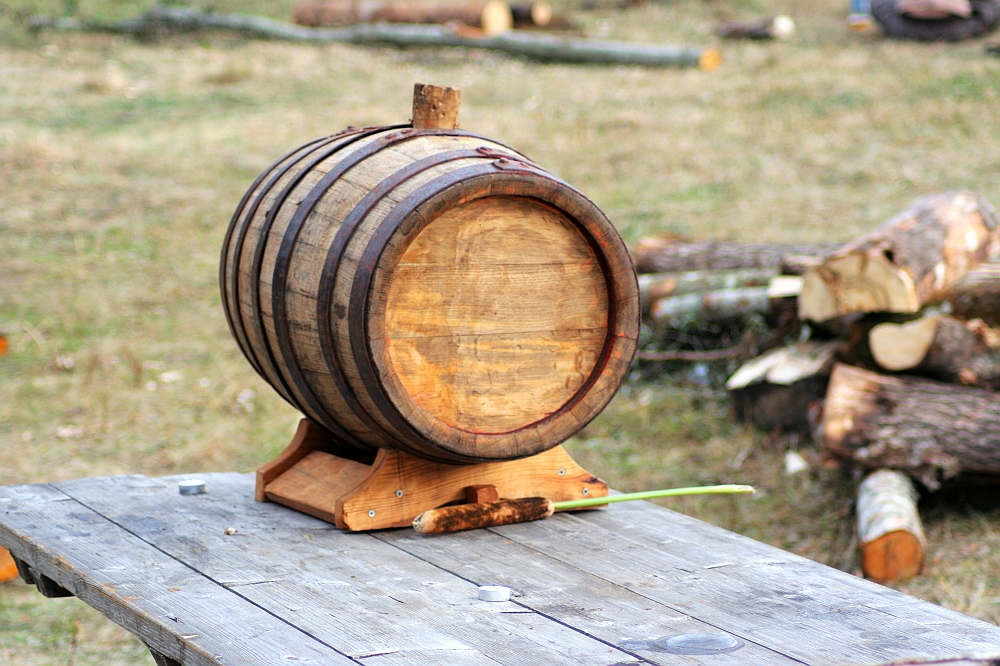 A small wooden barrel is placed on a wooden table outdoors. The barrel, featured in the 2008 event commemorating Semigallian days, is secured in a wooden stand and adorned with metal bands. In the background, there are chopped logs and rough wooden pieces scattered on the ground, indicating a rustic environment.