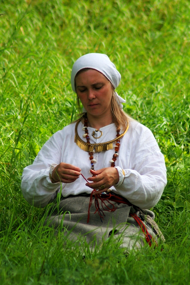 A woman in traditional Semigallian attire sits in a field of lush green grass. She wears a white headscarf, a white long-sleeved blouse, and a grey skirt. Focused on her task, possibly threading beads or weaving, she adorns multiple golden and beaded necklaces reminiscent of Semigallian days.