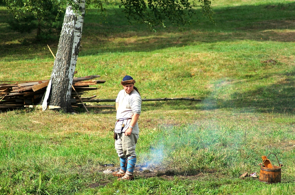 A person dressed in historical Semigallian clothing, including a tunic, belt, and leggings, stands on a grassy area. They are looking down at a spot with faint smoke visible. In the background, there is a stack of wood and a large tree. A wooden bucket and some utensils lie on the grass nearby.