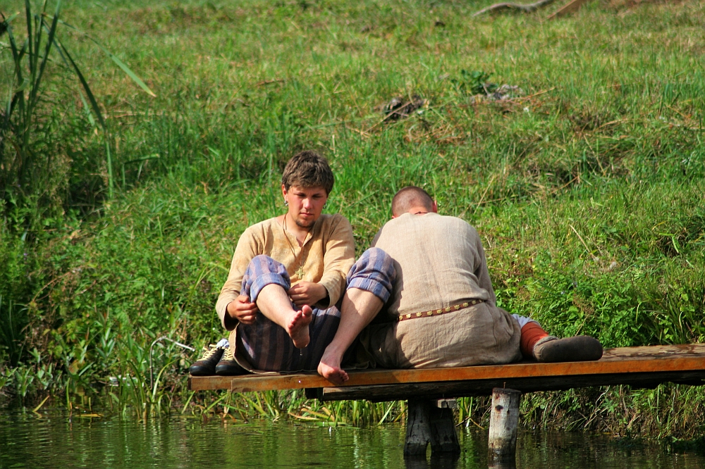 Amid the green grass and slightly sloped, natural landscape, two individuals clad in simple, weathered clothing sit on a wooden platform by the calm water's edge. The scene, reminiscent of Semigallian days past even in 2008, features one person tending to their feet while the other gazes into the serene waters.