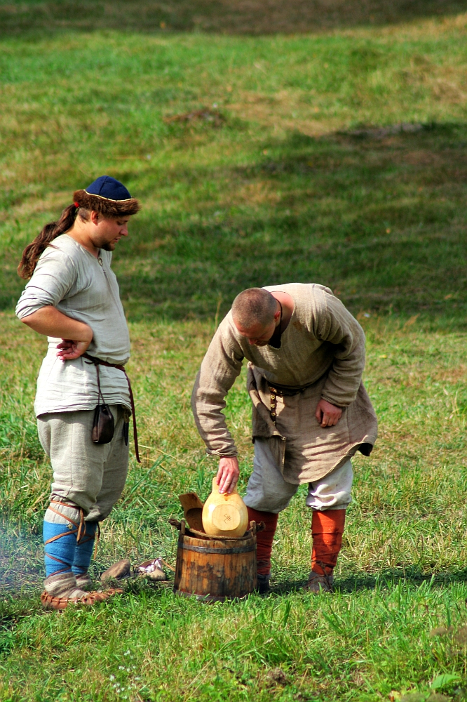 At the Semigallian festival, two people dressed in medieval-style clothing are in a grassy outdoor setting. One person, with long hair and a blue cap, stands with hands on hips, watching the other, who is bald and bent over, placing a round loaf of bread into a wooden barrel. They both wear tunics, trousers, and boots.