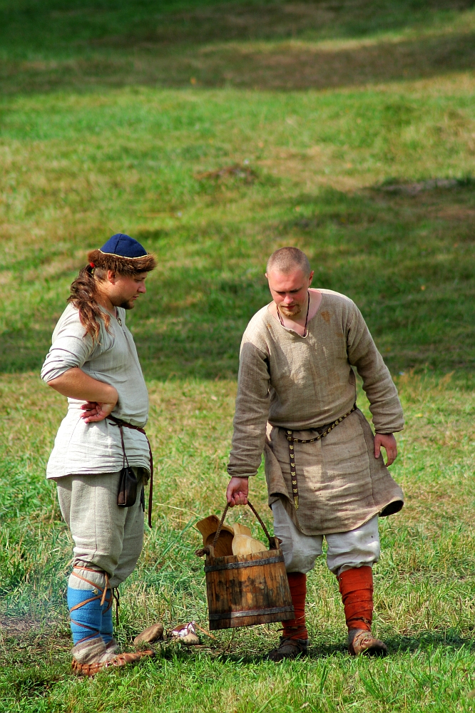 Two people wearing medieval-style clothing are standing on grassy terrain. One, with long hair and a headband, is watching the other, who is bald and holding a bucket by its wooden handle. Both wear neutral-toned tunics, belts, and colorful leg wraps amidst a green, sunlit backdrop reminiscent of Semigallian Days 2008.
