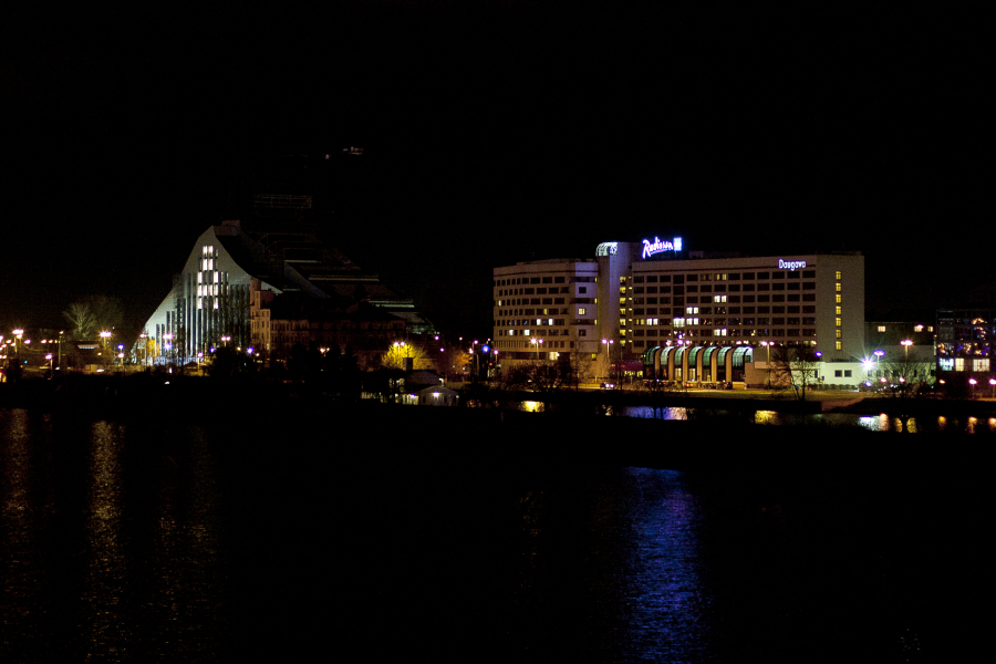 Nighttime image of a cityscape featuring two prominent, illuminated buildings by a body of water. On the left, the building has a modern, triangular design. On the right, a hotel with visible signage. Reflections of the lights shimmer on the dark water's surface during Staro Rīga 2011 festivities.
