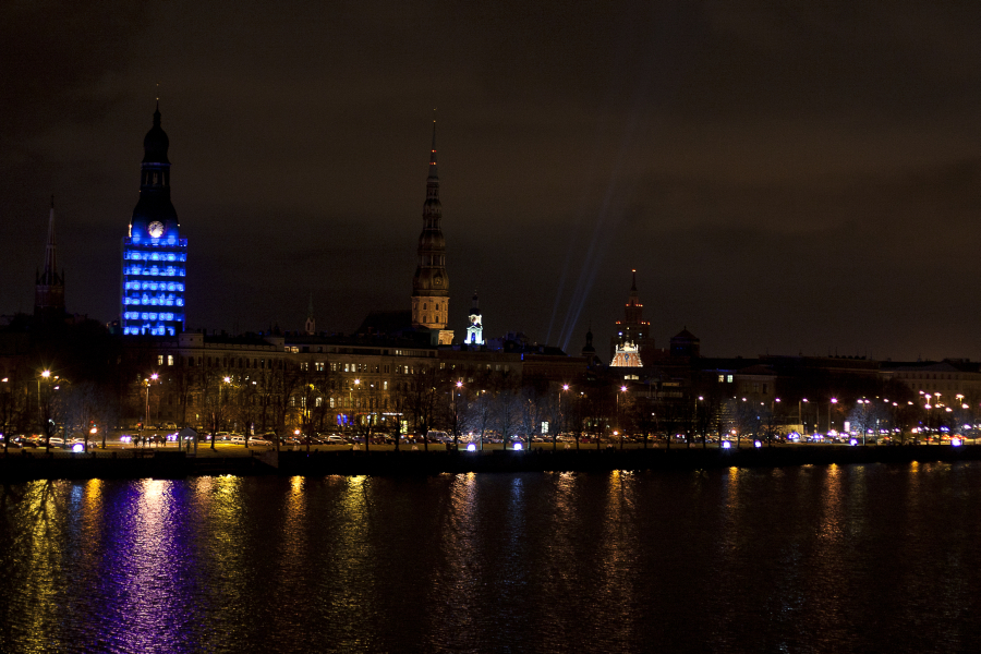 A nighttime cityscape featuring a skyline with various illuminated towers and spires showcases the charm of Staro Rīga 2011. A large building on the left glows blue, casting reflections on a river in the foreground. The dark sky is dotted with scattered clouds, while streetlights line the riverside, illuminating the scene below.