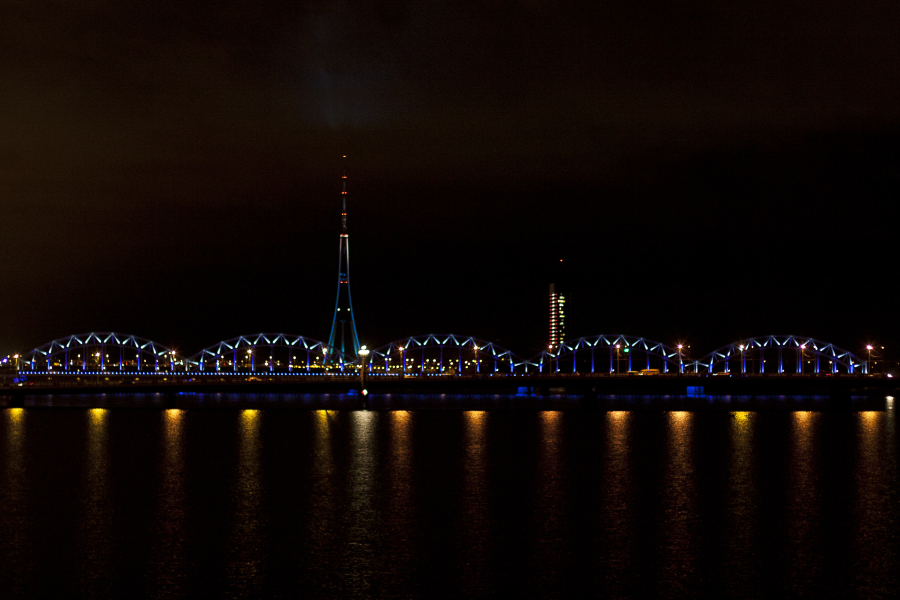 A nighttime view of the city skyline during the 2011 Staro Rīga festival features a brightly lit bridge spanning a river, with arched sections illuminated in blue and yellow lights. A tall tower with red lights at the top stands prominently in the background, and city buildings with lights on are visible to the right.