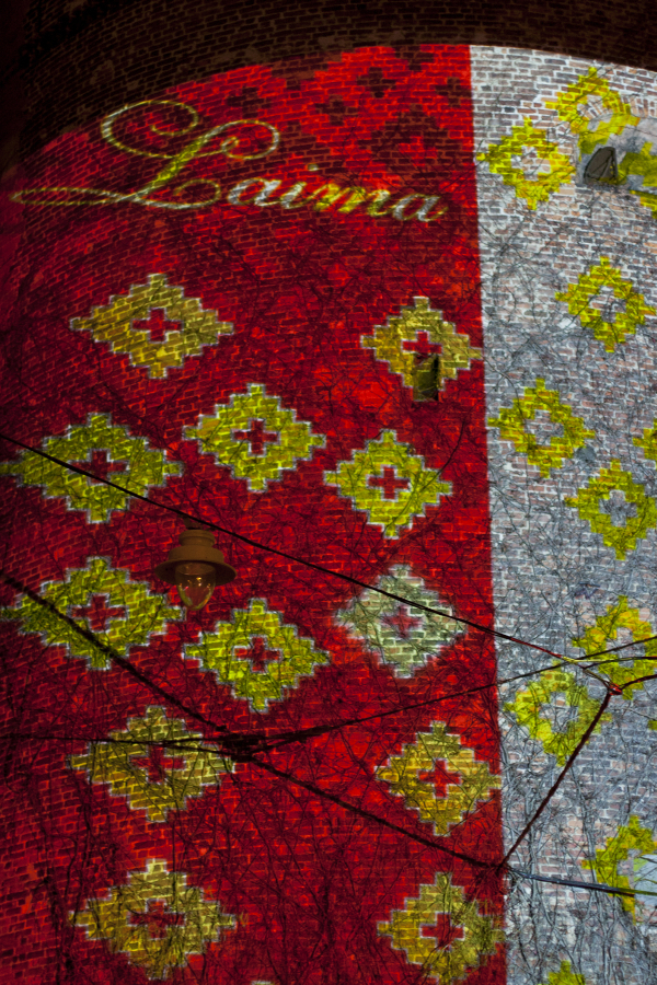 A brightly illuminated wall at the 2011 Staro Rīga festival displays a colorful pattern with red and yellow diamond shapes and an ornate script reading “Laima” at the top. Part of the wall is covered in brickwork, while the other part has a grayish texture. Vines and light fixtures are visible against the lit background.