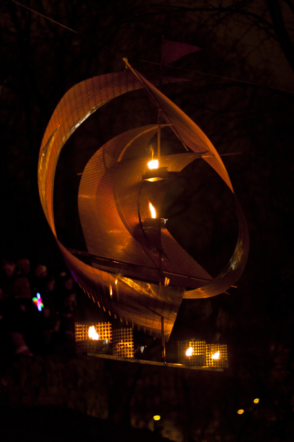 A metal sail-like sculpture illuminated by small lanterns is pictured at night. The abstract structure features curved metal sheets forming a spiral with glowing flames inside. In the background, silhouettes of people holding colorful lights are visible against a dark, blurry forest backdrop at Staro Rīga 2011.