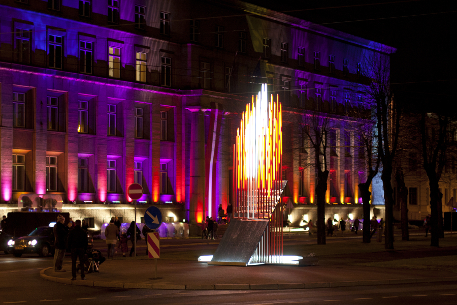 A nighttime scene from 2011 features a tall building illuminated by vibrant pink and purple lights. In front of the building is a modern art installation with vertical, illuminated rods in varying heights, resembling flames. The surrounding area is busy with people and vehicles, and colorful street signs are visible. This scene is part of the Staro Rīga festival.