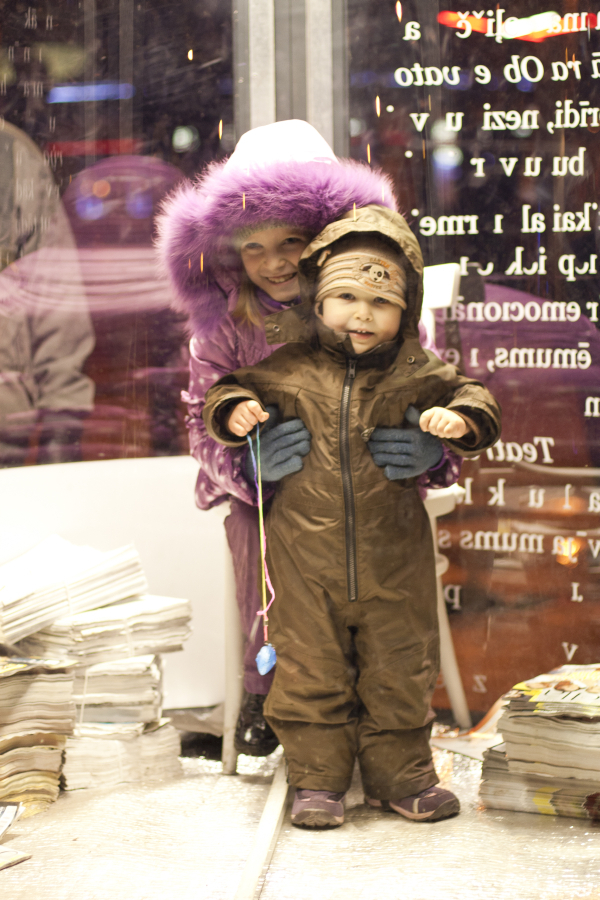 Two young children stand inside a brightly lit space with large windows. The older child, wearing a purple hooded coat, smiles and stands behind a smaller child in a brown snowsuit holding a toy on a string. Stacks of newspapers lie on the floor nearby. Snowflakes from the 2011 Staro Rīga festival can be seen falling outside.