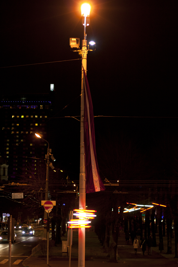 A streetlight illuminates the dark street with a warm, amber glow. A multicolored flag hangs from the pole. Below, traffic trails captured in long exposure appear as bright, colorful streaks. In the background, buildings and additional lights are visible against the night sky of Staro Rīga 2011.