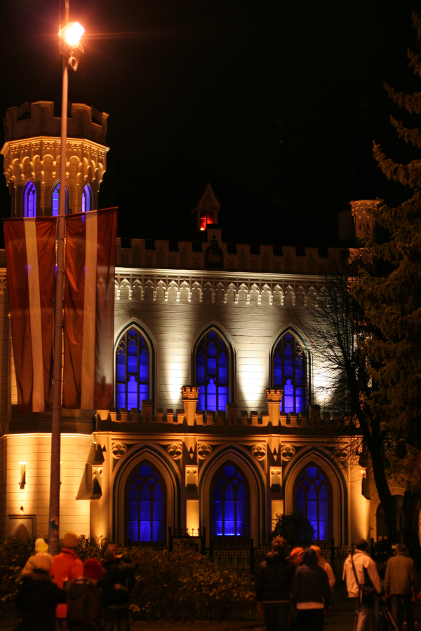 A nighttime image of a historic Gothic-style building with arched windows illuminated by blue and white lights during Staro Rīga 2011. A large pole with a red and white banner is in the foreground. Several people in winter clothing are gathered near the building, and the scene is adorned with a nearby tree.