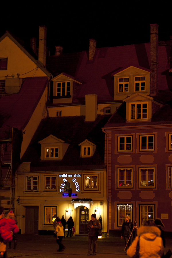 A nighttime street scene shows people walking past a row of charming, old-fashioned buildings with lit windows. One building features a brightly illuminated sign in blue with white text. The atmosphere, reminiscent of Staro Rīga 2011, is warm and inviting, with light spilling onto the cobblestone street.