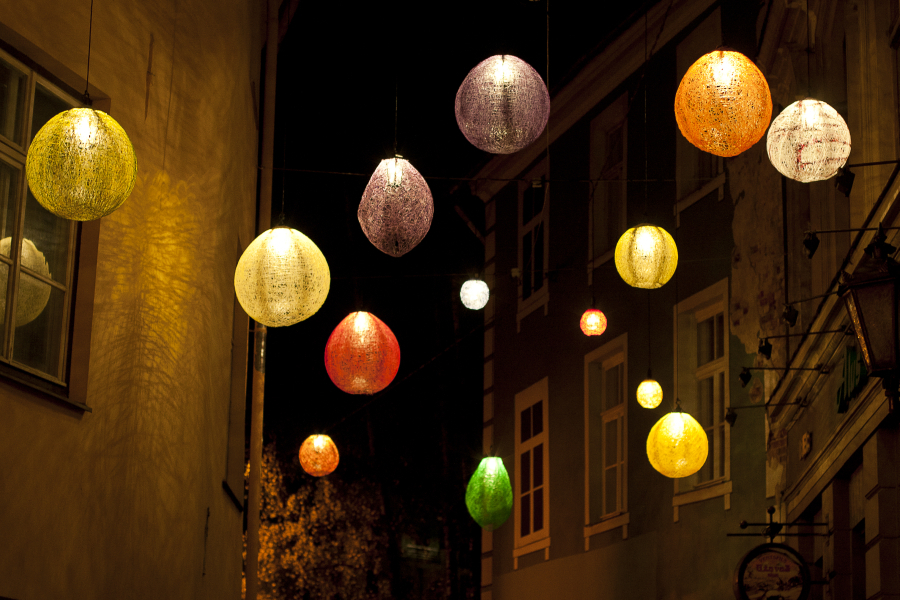 Colorful spherical lanterns in shades of yellow, purple, red, green, and orange hang above a narrow street at night during the 2011 Staro Rīga festival. The lanterns illuminate the area with a soft, warm glow against the backdrop of dark buildings. The scene evokes a festive and cozy ambiance.