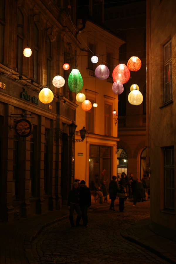 A warmly lit narrow street at night, featuring hanging colorful lanterns above. People walk on the cobblestone pathway lined with old buildings and shops. The atmosphere is cozy and inviting, with the soft glow of lanterns illuminating the scene—a snapshot reminiscent of Staro Rīga in 2011.