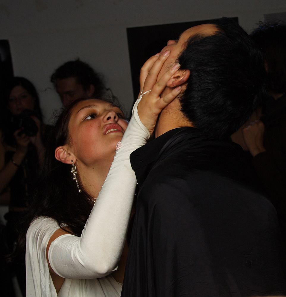 A woman wearing a white outfit and long white gloves is holding the face of a man dressed in black. Her expression is intense, her fingers pressing into his cheeks as she leans towards him, capturing a moment of raw emotion akin to māksla. The background is dimly lit with a few indistinct people visible.