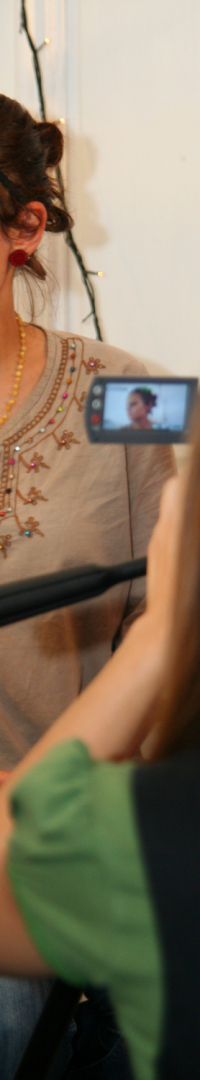 A person with a floral embroidered blouse sits on a stool, facing slightly to the left. Another person wearing a green T-shirt films them using a handheld video camera. String lights hang in the background, and the setting appears to be indoors with a casual preparty atmosphere.