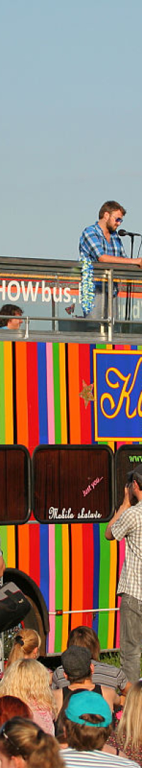 A large, colorful double-decker bus with red, yellow, and green stripes is parked outdoors, serving as a stage. Several people, including a cameraman, are on the top deck. A crowd of spectators is gathered around the bus at Oligarhu kapusvētki, with some taking pictures and videos. Signs read "Karneval" on the bus.