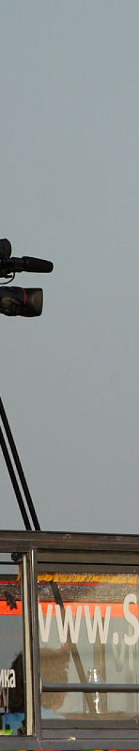 A person in a white shirt speaks into two microphones on an open-top bus during the kapusvētki festival. Another individual in a red shirt stands next to a video camera on a tripod, partially shaded by a red umbrella. The side of the bus displays the website "SHOWbus," and the sky is clear and blue in the background.